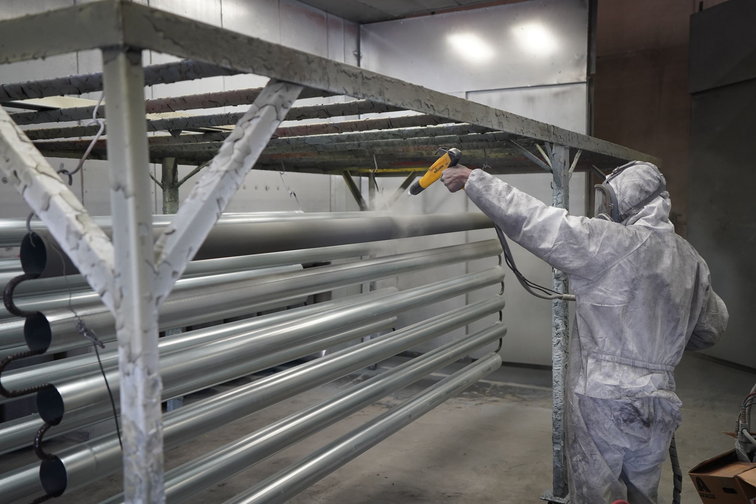 Worker in protective gear applying powder coating to metal pipes in a factory setting.